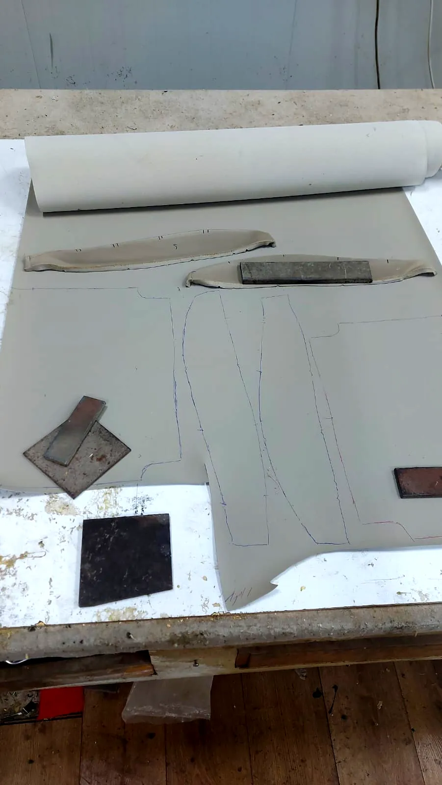 גליל חומר חדש פרוש על שולחן העבודה עם קווי תבנית מסומנים, לצד חלקים מקוריים ומשקולות מתכת
